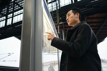 Young stylish Asian businessman in wireless earphones intently watching timetable of public transport at station