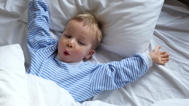 Cute funny cheerful little child boy in blue pajamas wakes up in the morning in bed, stretches and hides under the blanket again.
