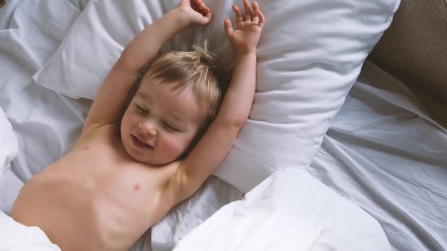 Cute funny little child wakes up in the morning in bed, stretches and hides under the blanket   again.
