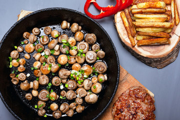 golden mushrooms champignon sprinkled with green onions in a cast iron black pan fries and juicy meat cutlet on forest wooden coasters on a gray background top view close-up