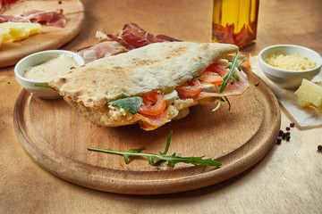 Close up view on tasty traditional italian sandwich panuozzo - oven-baked tortilla with tomatoes, mozzarella, ham and arugula on a wooden board. Street food