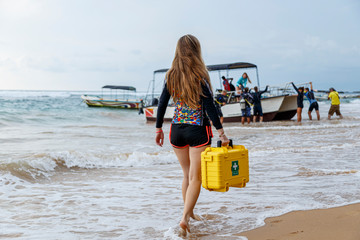 young blonde girl in diving suit carries a medical case with first aid to the boat, which is pushed out by a group of divers.