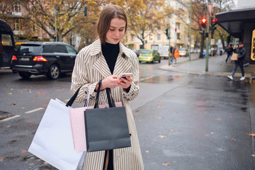 Attractive stylish girl with shopping bags happily using smartphone while standing on cozy city...