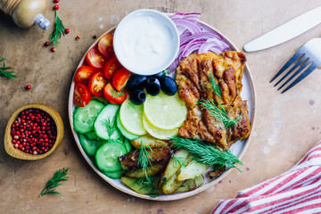 Lunch bowl with grilled chicken, red onion, tomato, cucumber, lemon, olive and sause on wooden background. Top view.