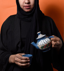 Arabic girl wearing Hijab pouring Tea into tea cup over on orange  background , girl in Arabic...