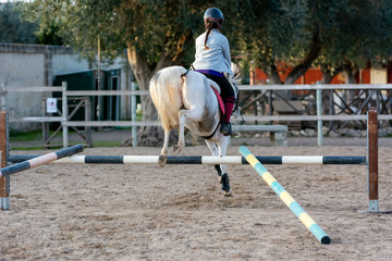 Back side of Girl riding a white horse jumping a guided obstacle during equestrian school training