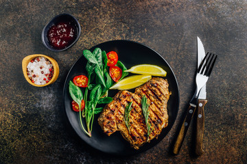 Roasted pork steak on black plate over dark background. With lemon, Cranberry Sauce and wine.