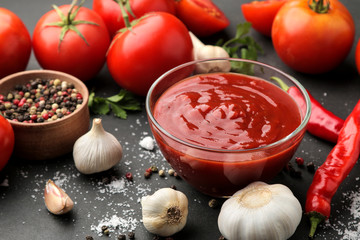 Red sauce or ketchup in a bowl and ingredients for cooking, spices, garlic, tomatoes and herbs on a black background.