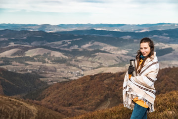 Naklejka premium Scenery autumn sunny day. Woman standing covered in plaid with termo cup in hand watching the mountains peak, sky with clouds.