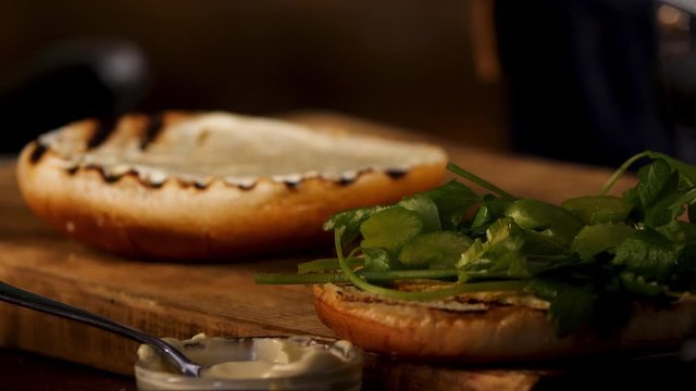 Craft burger is being cooked on dark room background. Stock footage. Close up of white sour cream sauce on a toasted burger bun lying on a wooden board near greenery.