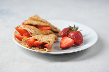 Thin pancakes with slices of strawberries.  A few strawberries on a plate next to pancakes.  Close-up.  Light background.
