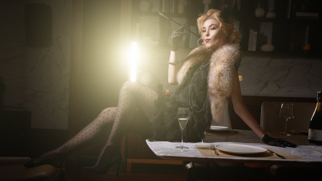 1920s retro style. Beautiful young woman in a dress sits on a table and smokes in a luxury interior, drinks champagne in a beautiful light. The photo shows soft noise for a vintage style. Color photo