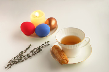 Easter colored eggs lie on a saucer next to a burning candle. White Cup with tea and cookies on a saucer Selective focus.