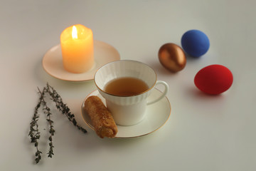 Obraz na płótnie Canvas Easter colored eggs lie on a saucer next to a burning candle. White Cup with tea and cookies on a saucer Selective focus.