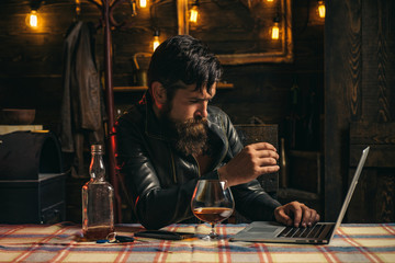 Stylish handsome bearded man enjoying whiskey and doing his business at laptop. Elegant man in leather jacket drinking a whiskey in bar. Brutal hipster man drinking alcohol.