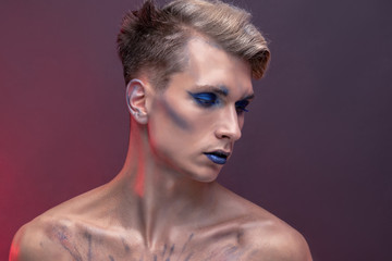 Portrait of a young man with professional beauty makeup and blond flying hair on a red background, beautiful eyes and lips with blue eyeshadows and lipstick, natural clean skin, face and hair care