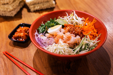 Shrimp Bibimbap Noodles, Korean noodles mixed with radish, sprout, carrot, cabbage, cucumber,  boil egg and Gochujang in red bowl served with Kimchi and red chopsticks. Perfect mouth watering cuisine.