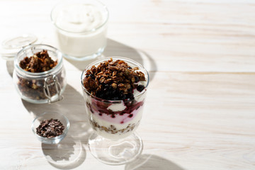 Yogurt parfait with cocoa granola, frozen blueberries and yogurt in bokeh. White wooden table, hard light, high resolution