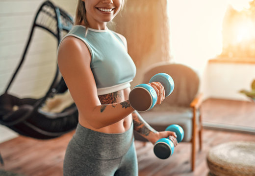 Cropped image of determined woman losing weight at home and exercising with dumbbells. Sport and recreation concept. Beautiful woman in sportswear with blue dumbbells in her hands.