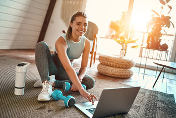 A sporty woman in sportswear is sitting on the floor with dumbbells and a protein shake or a bottle of water and is using a laptop at home in the living room. Sport and recreation concept.