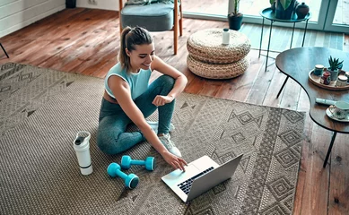 Poster A sporty woman in sportswear is sitting on the floor with dumbbells and a protein shake or a bottle of water and is using a laptop at home in the living room. Sport and recreation concept. © Valerii Apetroaiei