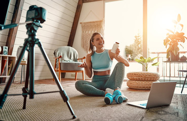 Woman blogger in sportswear sitting on the floor with dumbbells and a laptop and showing a jar of...