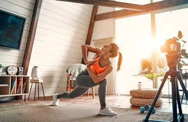 Athletic woman blogger in sportswear shoots video on camera as she does exercises at home in the living room. Sport and recreation concept. Healthy lifestyle.