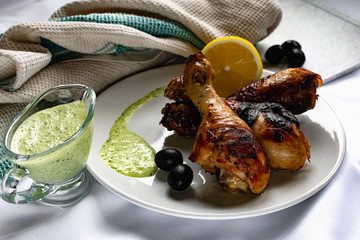 Toasted chicken drumsticks, with a brown Golden crust with fresh sauce with herbs decorated with lemon and black olives on a platter with a white textured napkin, towel
