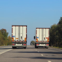 Two semi trucks with white van on suburban asphalt highway road at Sunny summer day against the blue sky and green forest on roadside, operative international cargo logistics spedition in Europe