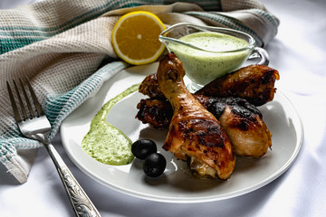 Toasted chicken drumsticks, with a brown Golden crust with fresh sauce with herbs decorated with lemon and black olives on a platter with a white textured napkin, towel