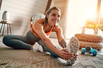 Fototapeta Athletic woman in sportswear doing fitness stretching exercises at home in the living room. Sport and recreation concept. obraz