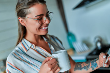A cheerful young woman in casual clothes and glasses holds a cup of hot coffee in her hand and writes a message on the phone against the background of a home kitchen interior.
