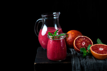 Smoothie with blood orange juice in the glass jar and jug on the edge of the wooden table. Still life against the black background