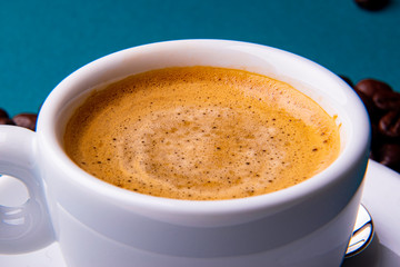 White espresso cup and saucer with brown coffee on a teal blue surface with beans and spoon.