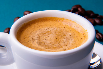 White espresso cup and saucer with brown coffee on a teal blue surface with beans and spoon.