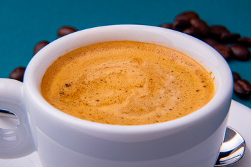 White espresso cup and saucer with brown coffee on a teal blue surface with beans and spoon.