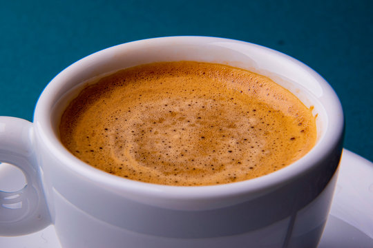 White espresso cup and saucer with brown coffee on a teal blue surface with beans and spoon.