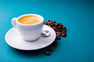 White espresso cup and saucer with brown coffee on a teal blue surface with beans and spoon.