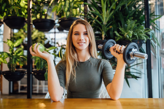 Portrait of pretty sporty girl sitting on modern gym, holding green apple, with dumbbells