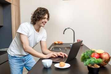 Portrait of handsome caucasian man, staying in casual white t-shirt use laptop in kitchen, healthy time concept