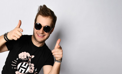 Male in sunglasses, black t-shirt, bracelets, jeans and sneakers. Smiling, showing thumbs up posing isolated on white. Full length