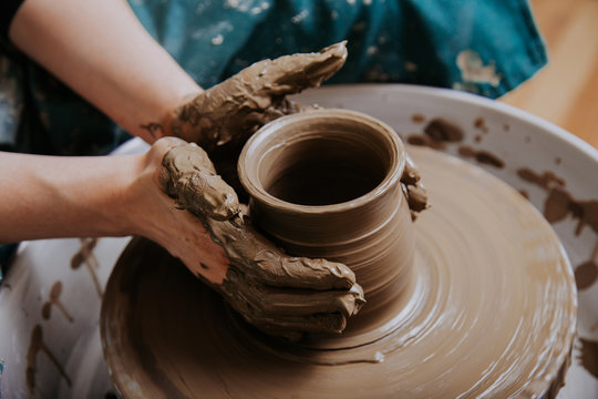 Close up mud covered hands of adult woman making clay pot on potter's wheel
