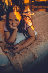 Obraz premium Young woman in home pajamas lying on bed at dawn. Adult lady resting, dangling legs and flipping through various news in mobile phone.