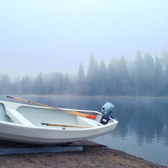 Small white boat with outboard engine and wooden oars laying on a shore at forest lake in Finland in a dense fog