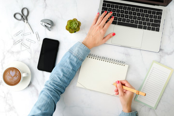 Flat lay workspace. Woman hand with coffee cup, smartphone, computer, notebook, planner and stationary with copy space on marble table background. Top view.