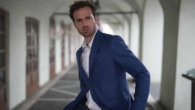 young businessman wearing navy suit walking with hands in pocket and then turning around holding hands on his jacket's button serious outside in the city