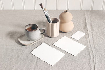 Artistic wokspace, still life. Paint brushes, pencils in ceramic holder, vase, cup of coffee and blank paper card mockups on linen tablecloth. Art supplies scene. Creative table background. Top view.