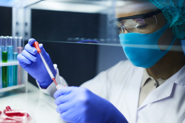Side view portrait of female scientist wearing mask and protective gear dropping blood samples in test tubes while working on research in laboratory, copy space