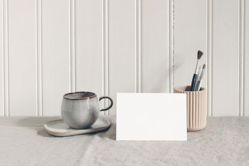 Blank paper card, invitatation mockup. Artistic stationery scene with paint brushes, pencils in ceramic holder and cup of coffee. Creative table composition, white wooden wall background.