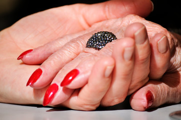 Adult daughter holding mother wrinkled old hand wearing a ring and red nail polish. Scene partially lit with a dramatic soft effect. Hands on the table/ adult daughter holding her mother hand/